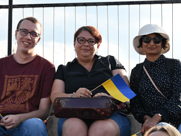 Ein Mann und zwei Frauen sitzen nebeneinander, die Frau in der Mitte hält eine Ukraine-Flagge in der Hand.